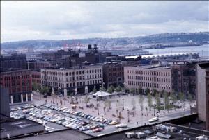 A city plaza surrounded by brick commercial buildings. In the distance is a body of water. The plaza has a few trees on it and there are several rows of parked cars next to it. 